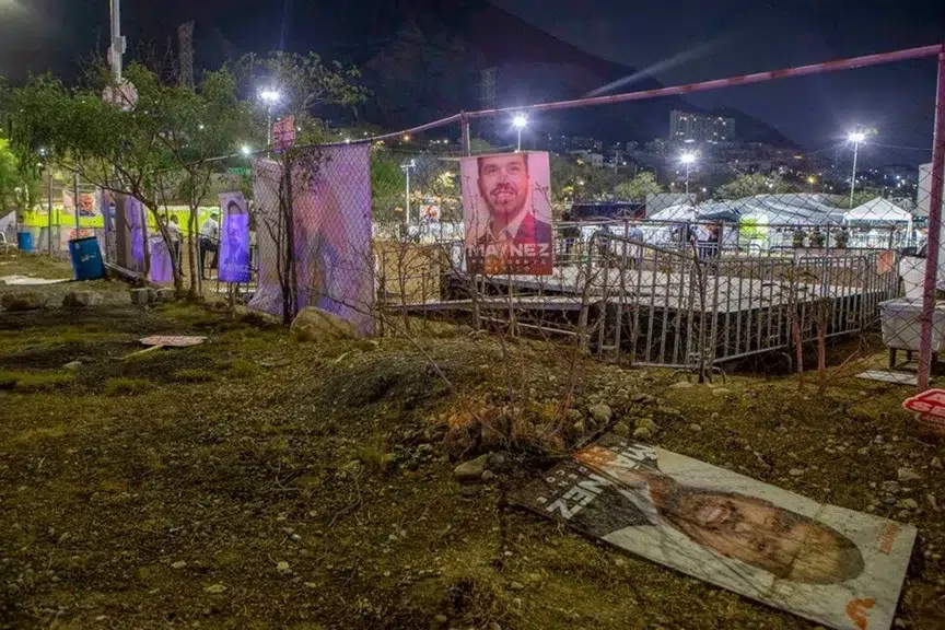 Una escena nocturna muestra una zona vallada adornada con carteles de campañas políticas, algunos tirados en el suelo, que recuerdan a debates recientes. Las luces iluminan el escenario con un podio, mientras que los árboles y edificios colocados contra un telón de fondo montañoso insinúan las noticias en México en desarrollo.