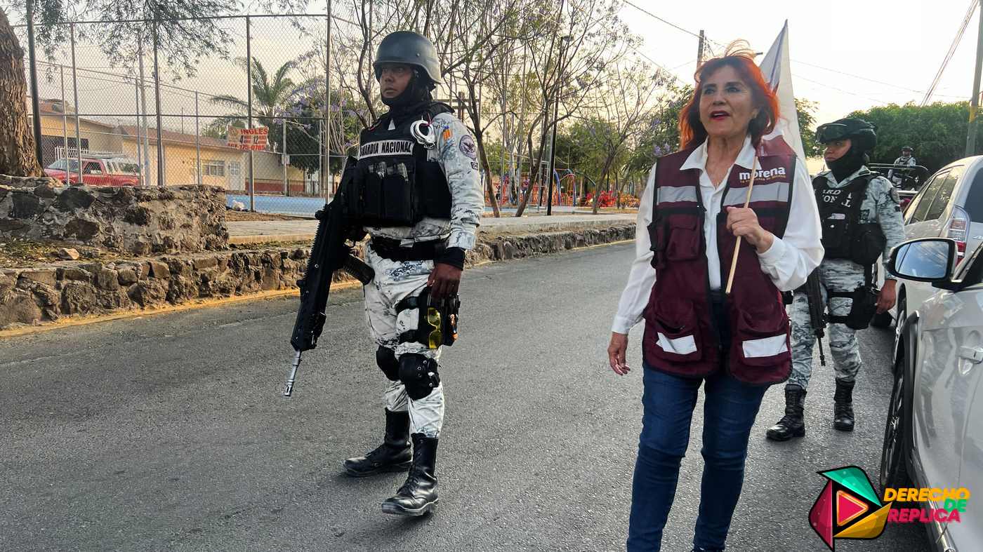 Una mujer con un chaleco marrón y blanco camina junto a dos agentes de seguridad armados con cascos y uniformes por una carretera pavimentada. Al fondo se ven árboles y edificios y se ve parcialmente un coche aparcado. El logotipo del "Derecho de Réplica" está en la esquina inferior derecha, destacando la preocupación por la violencia en las elecciones previas a las elecciones de 2024.