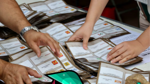 Varias manos están clasificando una colección de papeletas de votación extendidas sobre una mesa, cada papeleta muestra información impresa y logotipos diversos. La escena, que recuerda momentos cruciales en las democracias, sugiere un contexto de conteo u organización de votos en medio de posibles impugnaciones.